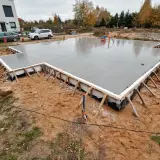 Świeżo wylany betonowy fundament, gotowy na budowę domu. Wokół plac budowy z piaskiem i trawą.