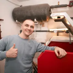 Uśmiechnięty mężczyzna pokazuje kciuk w górę obok czerwonego kotła i dużego zbiornika wyrównawczego CO.