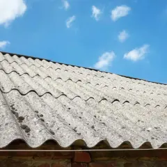 Stary dach pokryty eternitem, materiałem zawierającym azbest, pod błękitnym niebem z chmurami.