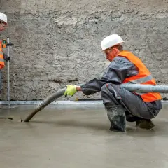 Dwóch budowlańców w kamizelkach odblaskowych wylewa beton. Jeden z nich kieruje wąż, z którego leje się masa, drugi kontroluje poziom. To praca z keramzytem budowlanym.