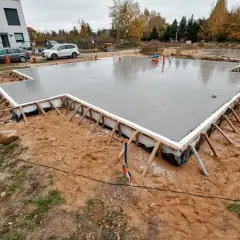 Świeżo wylany betonowy fundament, gotowy na budowę domu. Wokół plac budowy z piaskiem i trawą.