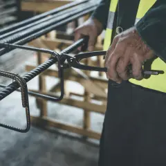 Ręce pracownika budowy zbroją konstrukcję z prętów ze stali węglowej, używając szczypiec do wiązania.