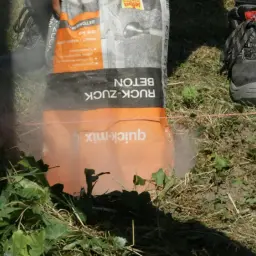 Szybki beton w worku "quick-mix" leży na ziemi obok buta roboczego. Idealny do szybkiego betonu.