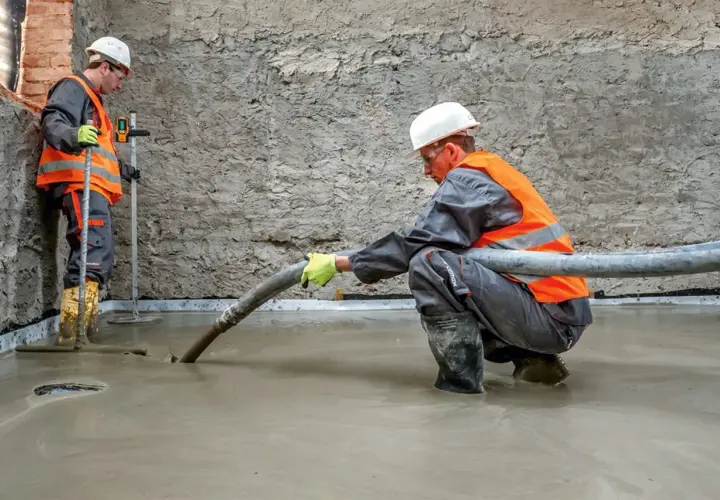 Dwóch budowlańców w kamizelkach odblaskowych wylewa beton. Jeden z nich kieruje wąż, z którego leje się masa, drugi kontroluje poziom. To praca z keramzytem budowlanym.