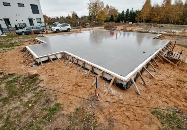 Świeżo wylany betonowy fundament, gotowy na budowę domu. Wokół plac budowy z piaskiem i trawą.