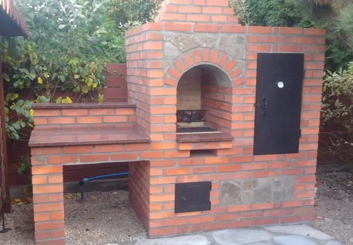 Solidny grill z cegły szamotowej, z kamiennym łukiem nad paleniskiem i metalowymi drzwiczkami. Idealny do ogrodu.