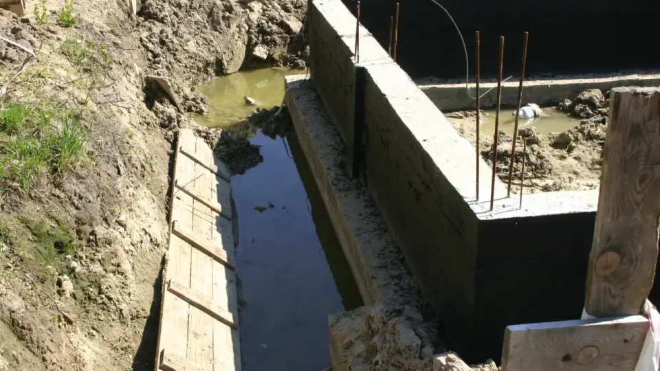Budowa fundamentów utrudniona przez wysoki poziom wód gruntowych. Wykop wypełniony wodą, widoczne zbrojenie i beton.