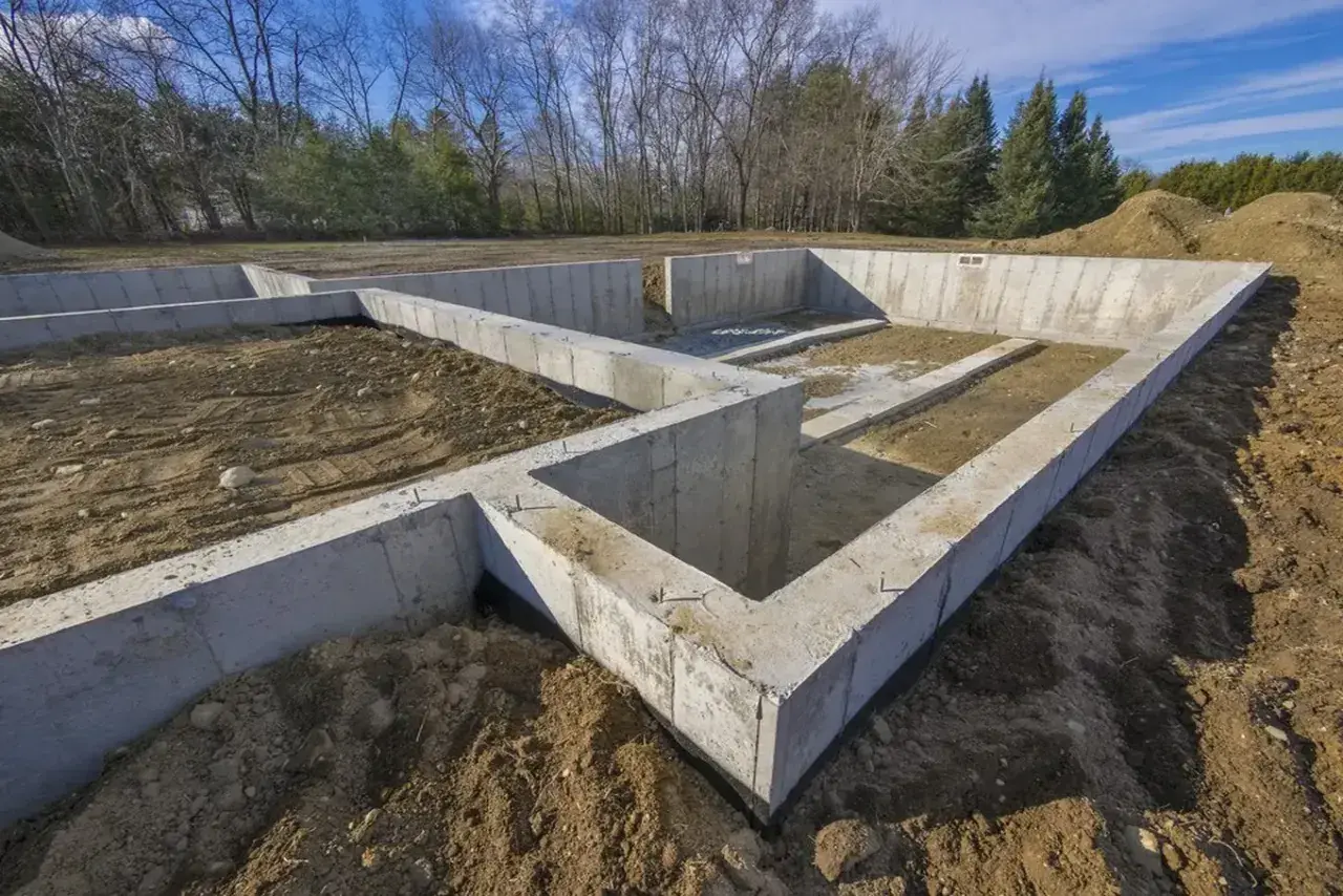 Solidny, jaki beton na fundament? Wylewane ławy fundamentowe pod przyszły dom, otoczone ziemią i drzewami.