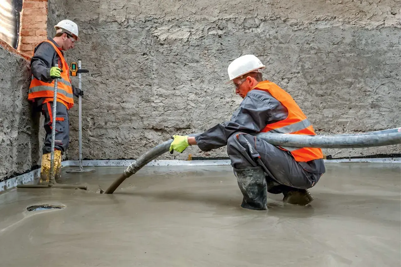 Dwóch budowlańców w kamizelkach odblaskowych wylewa beton. Jeden z nich kieruje wąż, z którego leje się masa, drugi kontroluje poziom. To praca z keramzytem budowlanym.