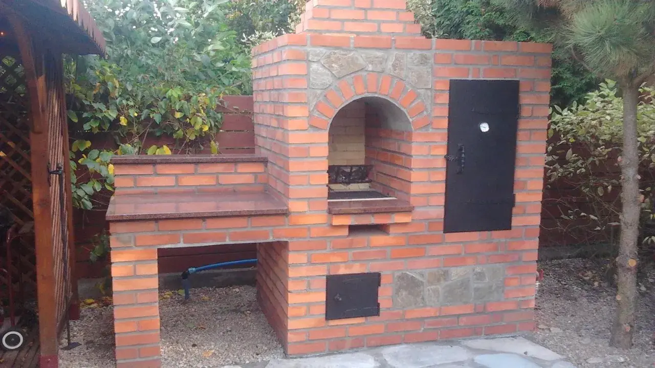 Solidny grill z cegły szamotowej, z kamiennym łukiem nad paleniskiem i metalowymi drzwiczkami. Idealny do ogrodu.
